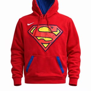 Puede incluir: Sudadera roja con capucha forrada en azul y un gran logotipo de Superman en la parte delantera. El logotipo es amarillo con un contorno negro y un aspecto desgastado. Un logotipo blanco de Nike es visible en la parte superior izquierda del pecho.