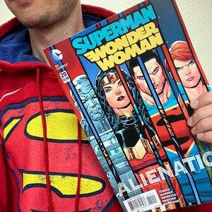Puede incluir: Una sudadera roja con el logo de Superman est&aacute; junto a un c&oacute;mic titulado "Superman Wonder Woman Alienation". La portada del c&oacute;mic presenta a Superman, Wonder Woman y otros personajes. El c&oacute;mic es el n&uacute;mero 20.