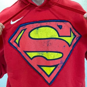 Puede incluir: Sudadera con capucha roja con el logotipo de Superman en amarillo, azul y rojo. El logotipo de Nike es visible en la parte superior izquierda. La capucha tiene un forro azul y cordones rojos.
