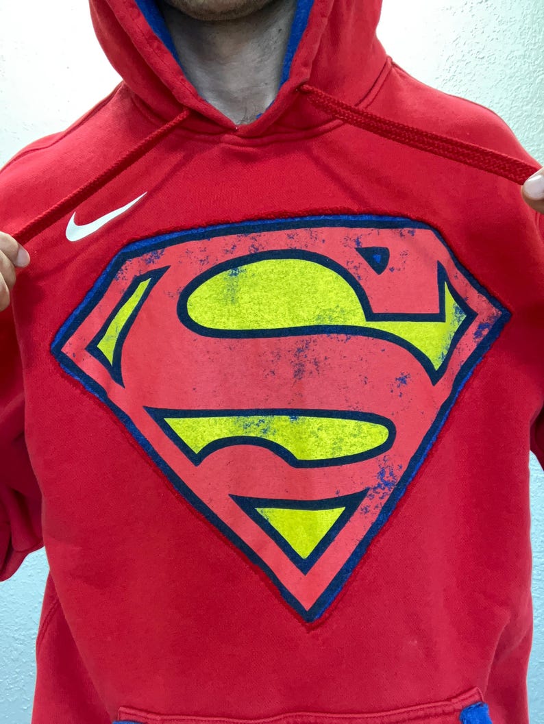 Puede incluir: Sudadera con capucha roja con el logotipo de Superman en amarillo, azul y rojo. El logotipo de Nike es visible en la parte superior izquierda. La capucha tiene un forro azul y cordones rojos.