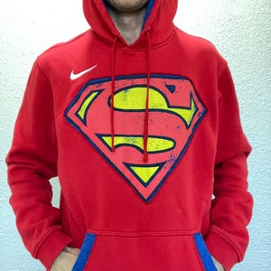Puede incluir: Sudadera con capucha roja con el logotipo de Superman en el centro. El logotipo tiene forma de escudo con una 'S' amarilla sobre fondo rojo, contorneada en azul. El logotipo de Nike est&aacute; en el pecho izquierdo. La capucha tiene forro azul.