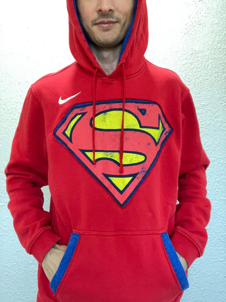Puede incluir: Sudadera con capucha roja con el logotipo de Superman en el centro. El logotipo tiene forma de escudo con una 'S' amarilla sobre fondo rojo, contorneada en azul. El logotipo de Nike est&aacute; en el pecho izquierdo. La capucha tiene forro azul.