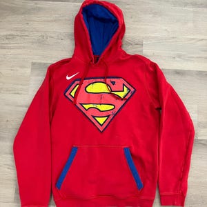 Puede incluir: Sudadera con capucha roja con interior azul y un gran logotipo de Superman en la parte delantera. El logotipo es amarillo, rojo y azul. Un logotipo blanco de Nike est&aacute; en el pecho superior izquierdo.