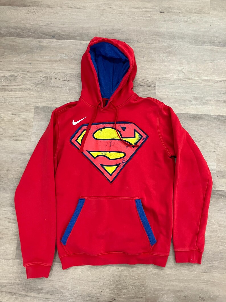 Puede incluir: Sudadera con capucha roja con interior azul y un gran logotipo de Superman en la parte delantera. El logotipo es amarillo, rojo y azul. Un logotipo blanco de Nike est&aacute; en el pecho superior izquierdo.