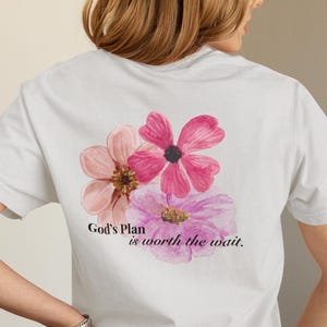 Puede incluir: Camiseta blanca con un diseño floral en acuarela en tonos rosas y morados. El texto "God's Plan is worth the wait" está impreso debajo de las flores. La camiseta es de estilo casual.