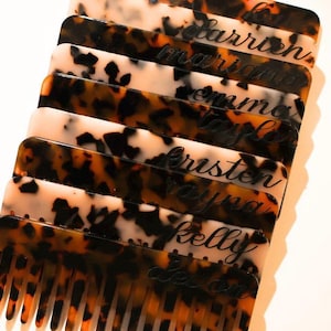 May include: A stack of seven rectangular hair combs featuring a tortoiseshell pattern in shades of brown, black, and cream. Each comb is engraved with a unique name in a cursive script, including the names Carrien, Mariana, Emma, Taylor, Kristen, Rayna, and Kelly.