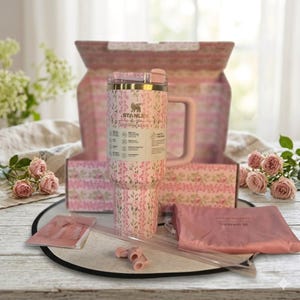 May include: A pink Stanley tumbler with a floral pattern and the text LoveShackFancy, featuring a handle and a clear straw. The tumbler is displayed in an open box with matching floral lining, accompanied by a pink pouch and small plastic components.