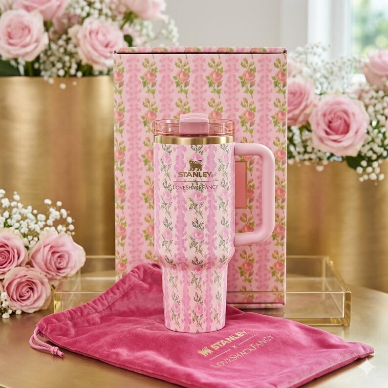 May include: A pink Stanley tumbler with a floral pattern, featuring a matching box and a dark pink velvet drawstring pouch. The tumbler has a handle, a lid, and the text Stanley and LoveShackFancy printed on the front and the velvet pouch.