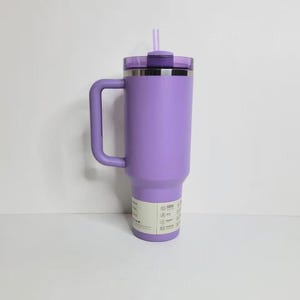 May include: A lavender-colored stainless steel tumbler with a large side handle, a matching purple lid, and a light purple straw. The cylindrical container features a white product information label wrapped around the lower section of the body.