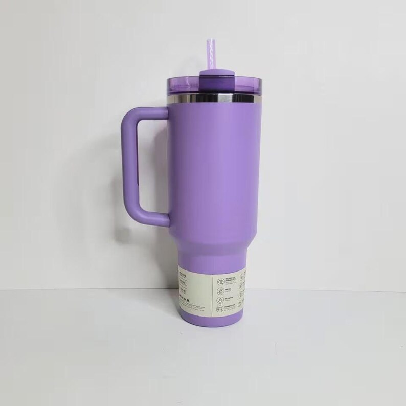 May include: A lavender-colored stainless steel tumbler with a large side handle, a matching purple lid, and a light purple straw. The cylindrical container features a white product information label wrapped around the lower section of the body.