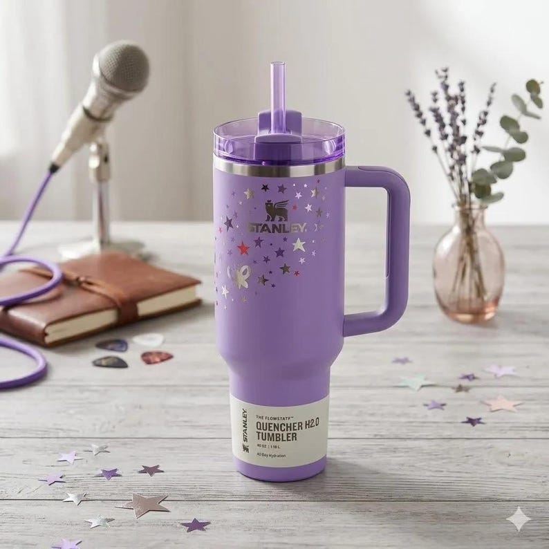 May include: A lavender Stanley Quencher H2.0 tumbler with a handle, a purple straw, and a clear lid. The body features a silver star pattern and the Stanley logo. A white label on the base reads The Flowstate Quencher H2.0 Tumbler 40 oz | 1.18 L.