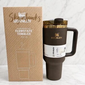 May include: A brown Stanley Quencher H2.0 FlowState tumbler with a 40 oz capacity and a matching handle, positioned next to its cardboard packaging box which features the text Skyhawks, Stanley, and The Quencher H2.0 FlowState Tumbler 40 oz 1.18 L.