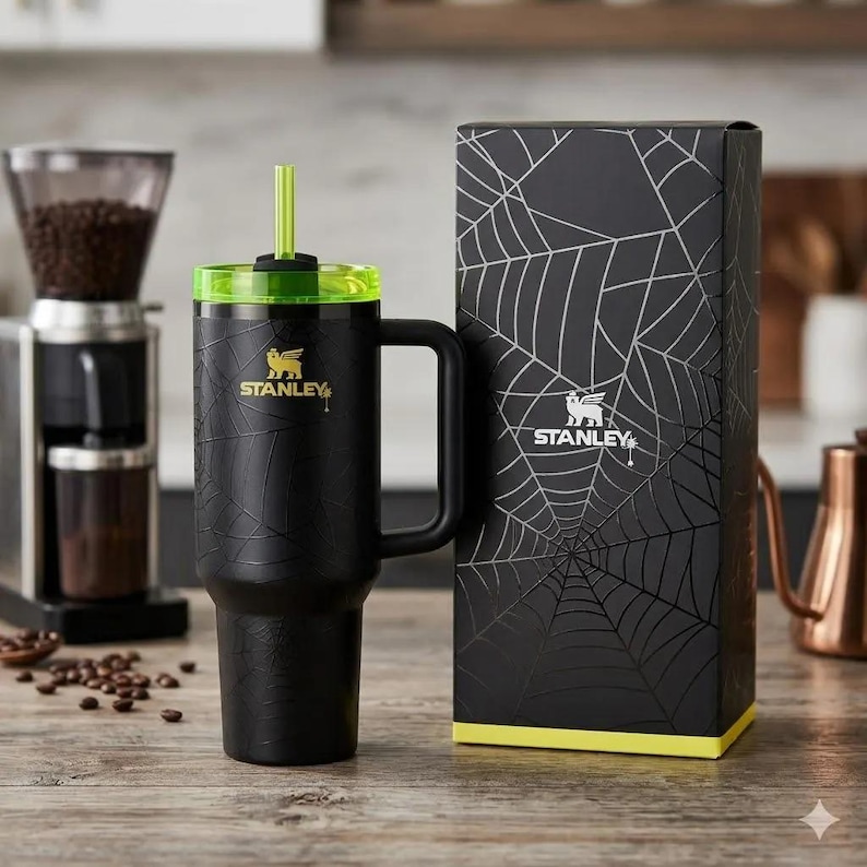 May include: A black Stanley tumbler with a spiderweb pattern, a lime green lid, and a matching straw. The tumbler features a gold Stanley logo and handle. It is displayed next to its matching black box, which also features a spiderweb design.