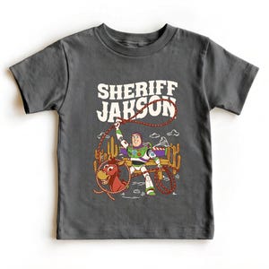 Personalized Sheriff Shirt, Custom Deputy Name T-Shirt, Space Ranger Cowboy Tee, Kids Western Birthday Gift, Toddler Buzz Lightyear Shirt

Step into an adventure-filled world for your little hero! This custom-designed t-shirt blends a beloved space-themed character with a fun western twist, sparking your child's imagination. It is the perfect outfit for birthday parties and a stylish choice for everyday playtime.