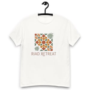 May include: A white short-sleeve t-shirt featuring a square graphic on the chest. The graphic displays a geometric tile pattern in shades of terracotta, sage green, and cream, accented by two palm leaves. The text RIAD RETREAT is printed below.