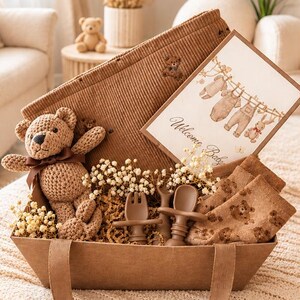 Op de afbeelding: Een bruine geschenkmand met een gehaakte teddybeer met een bruin lint, een bruine corduroy deken, een paar bruine sokken met patroon, een bruine siliconen lepel- en vorkset en een kaart met de tekst Welcome Baby en illustraties.