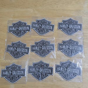 Może przedstawiać: Dziewięć naszywek Harley-Davidson. Każda naszywka przedstawia logo Harley-Davidson w kształcie tarczy, ze słowami "MOTOR HARLEY-DAVIDSON CYCLES" w czarno-białym wzorze. Naszywki są na przezroczystym podkładzie.