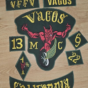 Puede incluir: Un conjunto de parches bordados con letras doradas sobre un fondo verde oscuro. El parche central representa una figura de diablo rojo con una rueda de motocicleta. Los parches incluyen las palabras "Vagos", "13", "6", "22", "1%", "California" y "VFFV".