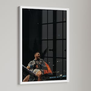 May include: A framed photograph of a man in a fighting arena. The man is wearing a patterned shirt, shorts, and a cape. The background is dark with a large window.