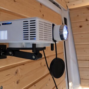 Op de afbeelding: Een witte projector gemonteerd op een zwarte verstelbare arm tegen een houten wand. De projector heeft een lens met helder blauw licht en een reeks verticale blauwe lichten aan de zijkant. Een zwarte kabel en lensdop hangen eronder.