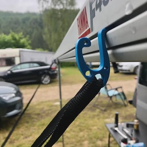 May include: A blue plastic hook attached to a white awning with the text "FIAMMA F45S" in red. A black strap is connected to the hook, with a blurred background of cars and trees.