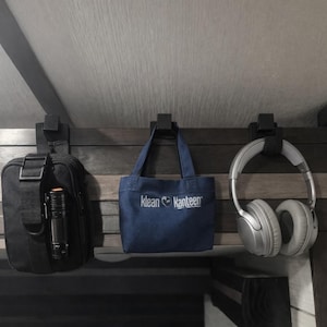 May include: A black bag with a flashlight, a blue tote bag with "Klean Kanteen" text, and silver headphones hanging from hooks on a wooden panel. The items are suspended from a dark-colored wooden structure.