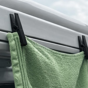Peut inclure: Gros plan sur une serviette verte maintenue par deux pinces à linge noires. Les pinces à linge sont fixées à une surface gris clair. La serviette est en tissu texturé.