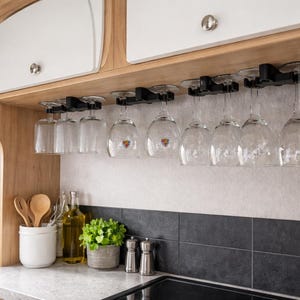 May include: A kitchen interior with a wooden cabinet and a row of wine glasses hanging upside down. The glasses are clear and have a stem. There are also wooden utensils in a white ceramic container, a small potted plant, and a black stovetop.
