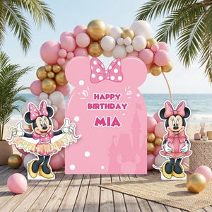 Puede incluir: Decoraci&oacute;n de fiesta de cumplea&ntilde;os con tem&aacute;tica de Minnie Mouse. Cuenta con un gran recorte rosa con el texto "Happy Birthday MIA", recortes de Minnie Mouse y un arco de globos en rosa, dorado y blanco. La escena est&aacute; ambientada en una playa.