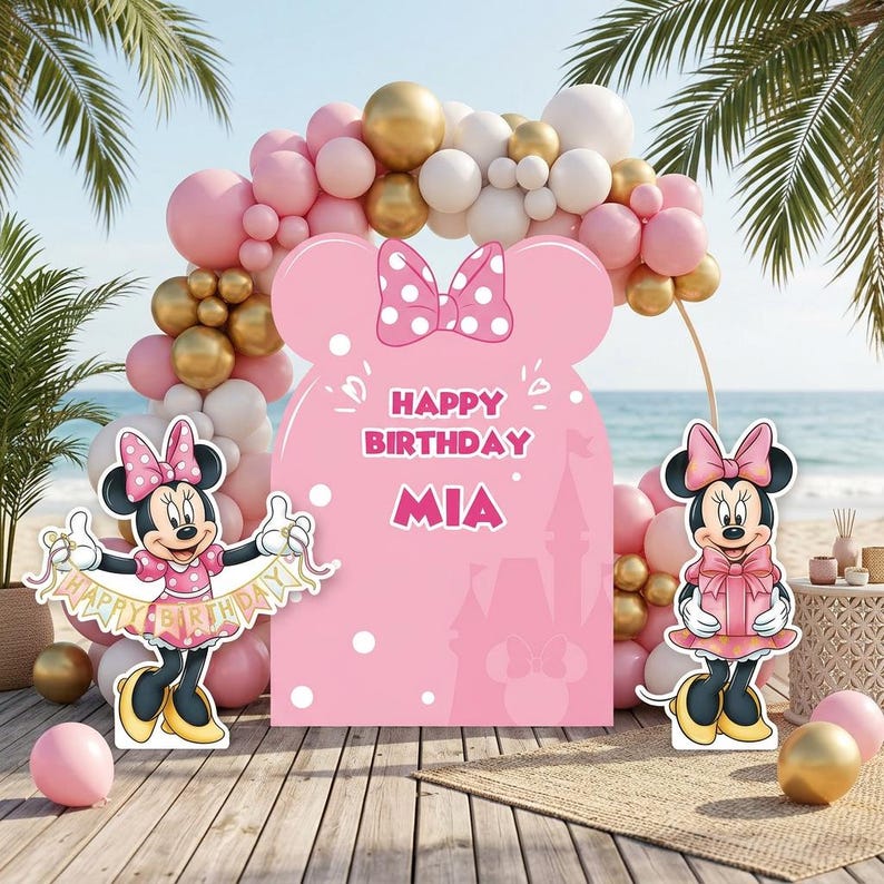 Puede incluir: Decoraci&oacute;n de fiesta de cumplea&ntilde;os con tem&aacute;tica de Minnie Mouse. Cuenta con un gran recorte rosa con el texto "Happy Birthday MIA", recortes de Minnie Mouse y un arco de globos en rosa, dorado y blanco. La escena est&aacute; ambientada en una playa.
