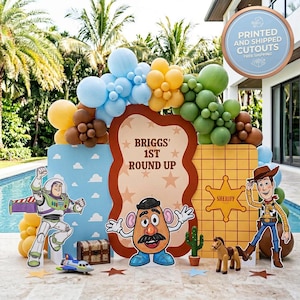 Puede incluir: Decoración festiva con recortes de personajes de Toy Story, como Buzz Lightyear, Woody y Mr. Potato. El fondo incluye globos y el texto "Briggs' 1st Round Up".