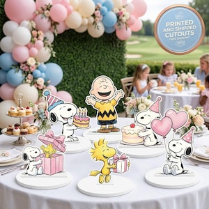 May include: A set of Peanuts character party cutouts featuring Snoopy, Charlie Brown, and Woodstock. The figures include Snoopy with a birthday cake, Snoopy with balloons, Snoopy with a gift, and Woodstock with a present. Each cutout is mounted on a white circular base.
