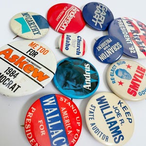May include: A collection of eleven round political campaign buttons in various colors including red, white, and blue. The buttons feature text such as McCarthy, Anderson for President, Gary Hart, Askew for President, Church for Idaho, Andrus, and Wallace.
