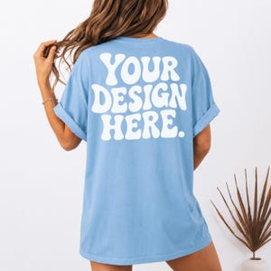 Op de afbeelding: Een lichtblauw T-shirt met korte mouwen en de witte tekst "YOUR DESIGN HERE." in een groot, vet, golvend lettertype op de achterkant. Het shirt heeft een ronde hals en opgerolde mouwen voor een ontspannen pasvorm.