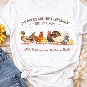 May include: A light gray t-shirt featuring a whimsical illustration of various fowl, including ducks, chickens, and a turkey. The text reads "MY DUCKS ARE MOST CERTAINLY NOT IN A ROW - AND I'M not even sure all of them are Ducks-".