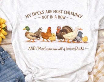 May include: A light gray t-shirt featuring a whimsical illustration of various fowl, including ducks, chickens, and a turkey. The text reads "MY DUCKS ARE MOST CERTAINLY NOT IN A ROW - AND I'M not even sure all of them are Ducks-".