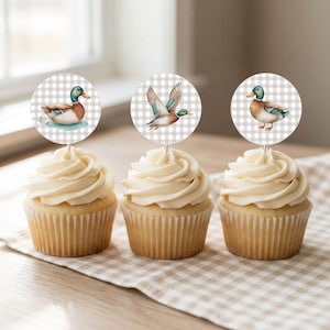 May include: Three cupcakes with white frosting, each topped with a round duck-themed image. The duck illustrations feature a mallard in various poses against a gray and white checkered background. The cupcakes sit on a checkered cloth.