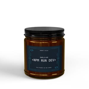 May include: A brown glass jar candle with a black lid. The label on the candle reads "Smells Like <NPM RUN DEV>" in white text on a dark blue background. The candle is an eco-friendly soy wax candle.