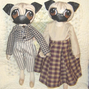 May include: Two handmade pug dolls with painted faces and fabric clothing. One doll wears a plaid shirt and striped trousers, the other a beige top and plaid skirt. The dolls are holding hands.