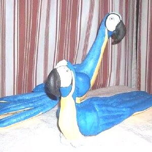 Puede incluir: Dos figuras de loros guacamayos azules y amarillos con picos negros y parches faciales blancos. Los loros están colocados con uno de pie y el otro acostado, ambos con alas azules texturizadas y detalles amarillos en el pecho.
