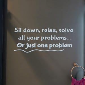 May include: A dark grey bathroom wall with white text saying "Sit down, relax, solve all your problems... Or just one problem." A purple towel hangs on a gold towel ring. A lit mirror is on the right.