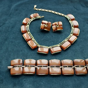 Könnte beinhalten: Ein Vintage-Schmuckset mit Halskette, Armband und passenden Ohrringen. Das Set hat einen goldfarbenen Metallrahmen mit rechteckigen, karamellfarbenen Akzenten. Der Schmuck wird auf einem dunkelblauen Stoff präsentiert.