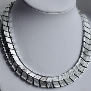 Könnte beinhalten: Eine silberfarbene Halskette bestehend aus einer Reihe miteinander verbundener, chevronförmiger Metallglieder. Jedes einzelne Glied weist eine texturierte, gebürstete Oberfläche auf, die ein mattes Erscheinungsbild über das gesamte Schmuckstück erzeugt.