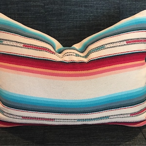 May include: A white pillow with colorful stripes in shades of red, pink, blue, and teal. The pillow is made of a woven fabric and has a slightly textured surface.