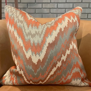 May include: Square decorative pillow with a woven geometric pattern in shades of orange, gray, and cream. The pillow is placed on a tan leather chair, with a brick wall in the background. The pillow's design features a zig-zag pattern.