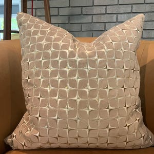 May include: A square decorative pillow with a repeating geometric pattern. The pillow has a light brown background with a cream-colored, four-petal design. The pillow is on a brown leather couch.