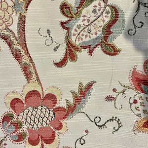 May include: Close-up of a floral fabric with a cream background. The design features large, stylized flowers and leaves in shades of red, green, and gold. The fabric has a textured appearance, with subtle vertical lines.