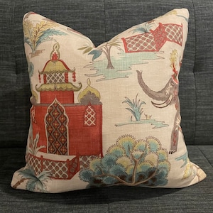 May include: A decorative throw pillow with a beige background and a colorful pattern of red, green, blue, and yellow. The pattern features an elephant, a building, and trees.