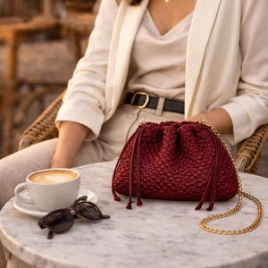 May include: A burgundy handbag with a gold chain strap sits on a marble table. The bag has a woven texture and drawstring closure. A cup of coffee and sunglasses are also on the table. The person is wearing a white blazer and beige pants.