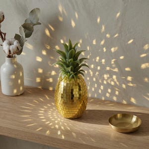 May include: A decorative pineapple sculpture with a gold mirrored surface and green leaves. The pineapple sits on a wooden shelf next to a small brass bowl and a white vase with cotton stems. The wall behind the pineapple is illuminated with a pattern of light.